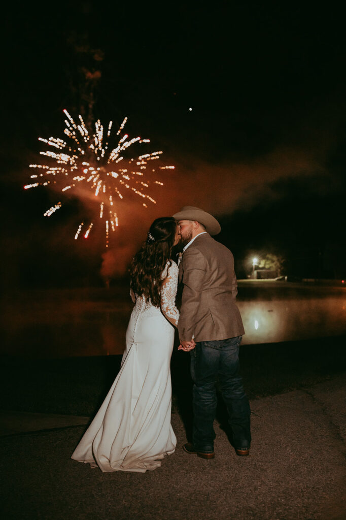 Bride and groom kissing after their wedding with a lake and fireworks as the backdrop